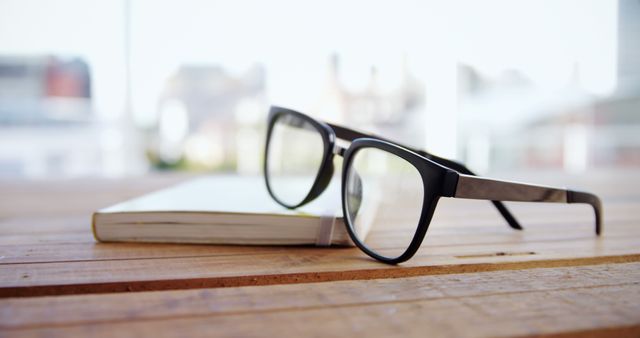 Black glasses resting on top of a closed notebook placed on a wooden table outdoors with blurred buildings in the background. Ideal for concepts related to reading, studying, relaxation, planning, eyewear, lunchtime breaks, optometry, and focus. Suitable for use in academic, optometry, productivity, and lifestyle websites or marketing materials.