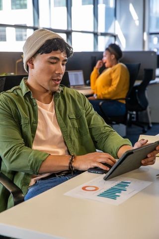 Asian man using tablet while coworker collaborating in modern open-plan office with charts
