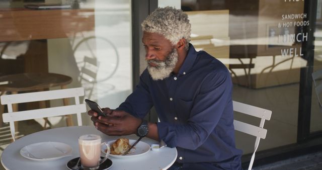 Senior man with gray hair and beard enjoying a coffee and pastry at an outdoor café while using his smartphone. Suitable for themes depicting leisure, technology use among older adults, lifestyle, relaxation, morning routines, and social interaction.