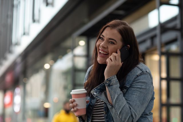 Curvy Caucasian woman enjoying a conversation on her smartphone while holding a takeaway coffee in the city. She is laughing happily, dressed casually in a denim jacket, with a modern building in the background. Perfect for use in advertisements, lifestyle blogs, social media posts, and articles about urban living, communication, and happiness.