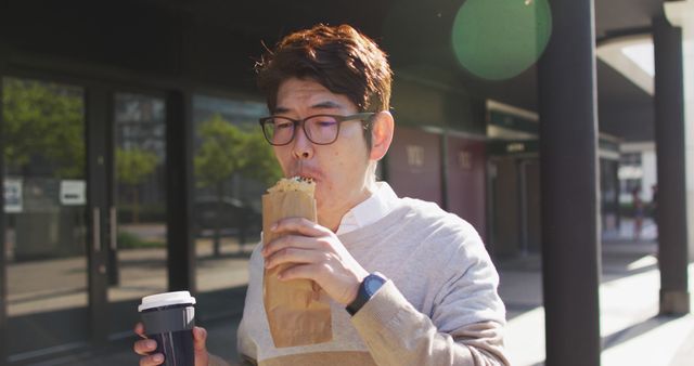 A man in casual attire and glasses enjoys sipping coffee and eating a pastry while walking on an urban street. This image is ideal for use in lifestyle blogs, morning routine articles, food and coffee advertisements, or fashion spreads that depict everyday life.