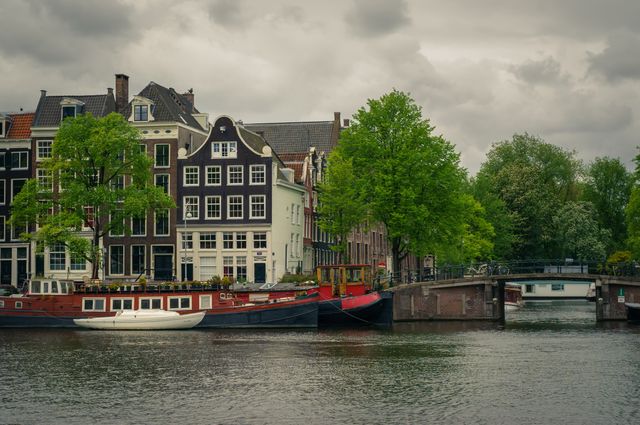 Charming scene featuring historical buildings and houseboats alongside an Amsterdam canal under a cloudy sky. Ideal for travel promotions, tourism websites, European architecture themes, and urban life concepts. Can be used to highlight city tourism, historical architecture, charming cityscapes, or writing backgrounds about urban development.
