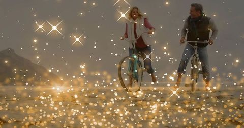 Mature couple riding vintage bikes along misty beach with golden bokeh lights