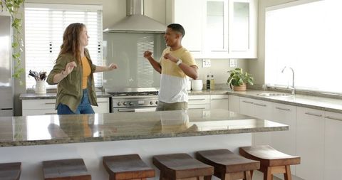 Young couple dancing playfully on sunlit modern kitchen with large granite island