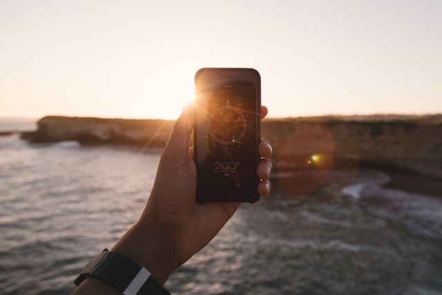 Hand holding smartphone displaying a compass app with the ocean and sun setting in the background. Ideal for themes related to travel, navigation, adventure, exploration, technology, and outdoor activities. Useful for travel blogs, tech tutorials, and adventure advertisements.