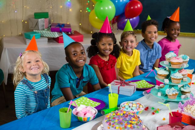 Children sitting at a table during a birthday party, wearing colorful party hats and surrounded by festive decorations. The table is filled with cupcakes, sweets, and gifts. Ideal for use in advertisements, party planning, educational materials, and family-oriented content.
