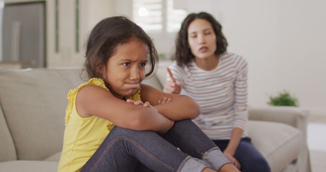 A young girl is sitting on a sofa with a look of frustration while her mother is scolding her from the background. This portrays scenes of parenting and disciplining within a home environment. The image can be used in articles or advertisements related to parenting challenges, family dynamics, behavior management, childhood education, and emotional development.