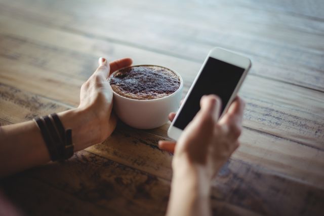 Woman holding a cup of coffee and using a smartphone at a wooden table in a cafe. Ideal for illustrating themes of modern lifestyle, technology use, relaxation, and coffee culture. Suitable for blogs, social media posts, and advertisements related to cafes, technology, and leisure activities.