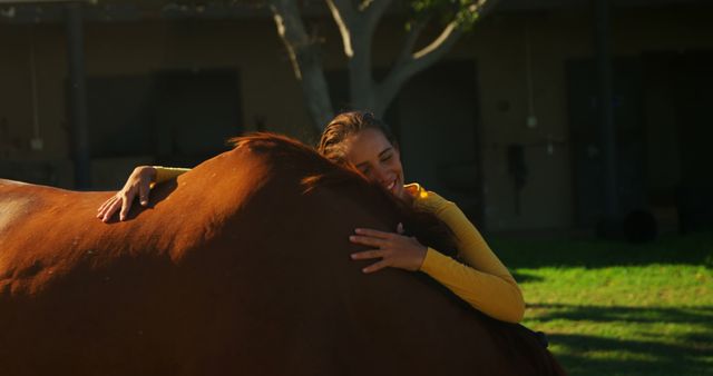 Young woman enthusiastically hugging a horse in a sunny outdoor setting. The background features greenery, suggesting a rural environment. The woman wears a yellow top, reflecting a cheerful and warm atmosphere. Great for illustrating themes of animal bonding, nature, and equestrian activities. Perfect for websites, articles, or advertisements related to pet care, outdoor activities, or mental wellness.