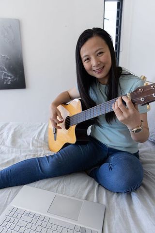 Asian woman playing acoustic guitar on bed with laptop, smiling and practicing music