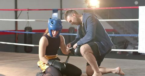 Coach adjusting female boxer headgear and gloves while kneeling in boxing ring
