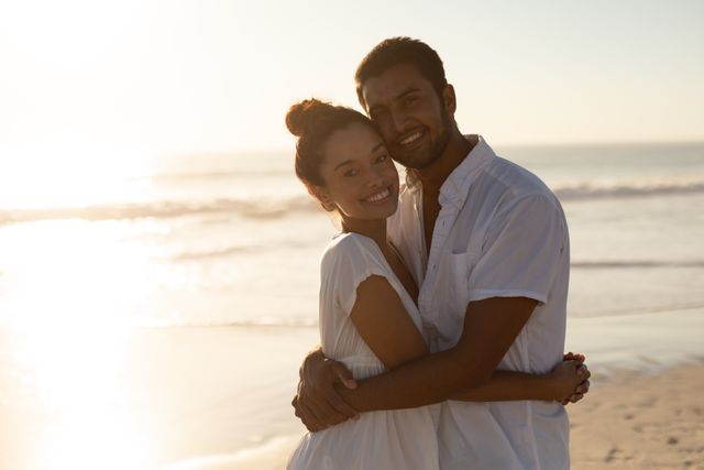Joyful couple embracing and smiling on a sunny beach at sunset. Perfect for illustrating romance, vacations, happiness, and relationships. Can be used for travel brochures, romantic-themed content, or lifestyle blogs.