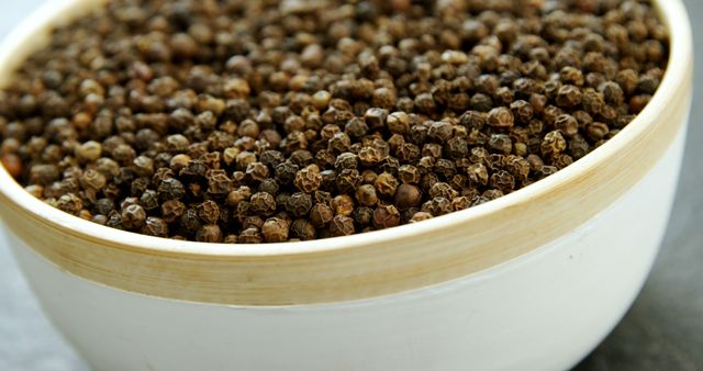 Close-up view of black peppercorns stored in a white bowl placed on a gray surface. Ideal for use in culinary articles, kitchen blogs, food preparation guides, or classes on organic and natural ingredients.