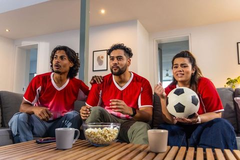 Tense asian football fans watching live match on couch wearing red jerseys with popcorn at home