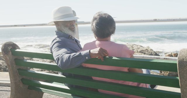 This image portrays a senior couple enjoying a relaxing moment on a green bench by the ocean. The man, with a beard and hat, places his arm over the back of the bench, while the woman sits beside him. This picture can be used for promoting retirement, senior living, travel, or leisure activities, highlighting the peaceful and joyful aspects of coastal living.
