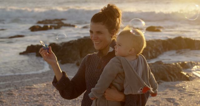 Happy caucasian mother blowing bubbles and carrying toddler son on beach at sunset. Motherhood, childhood, care, free time, travel, nature and healthy outdoor lifestyle, unaltered.