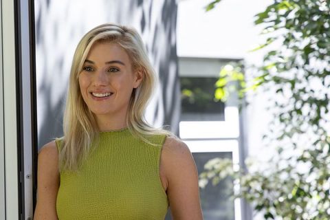 Smiling blonde woman leaning at doorway wearing green sleeveless top in sunlit garden