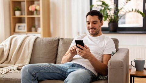 Smiling man using smartphone on sofa in cozy living room with plants and shelves