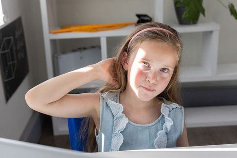 Preteen girl sitting at desk touching neck with sunlit patch on face in home study during morning