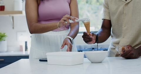 Diverse couple scooping vanilla ice cream into sugar cone on modern marble kitchen island