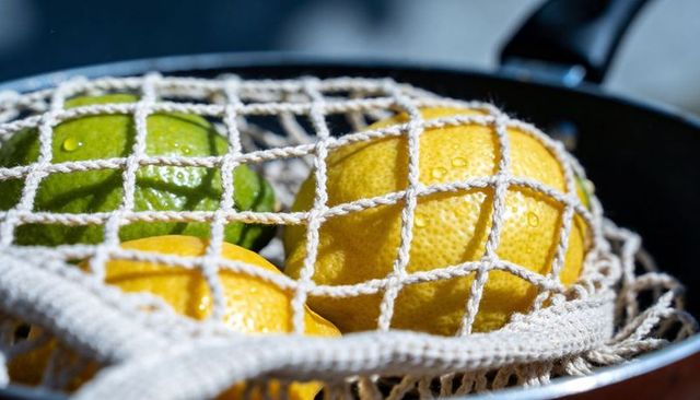 Dewy lemons and lime nesting in reusable mesh bag over colander rim in kitchen
