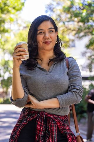 Young woman holding coffee on sunlit campus walkway, plaid shirt tied at waist