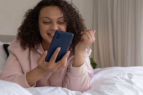 African american woman relaxing on bed smiling while using smartphone for messaging