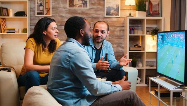 Friends watching soccer on tv, holding beer and sharing popcorn on cozy living room sofa