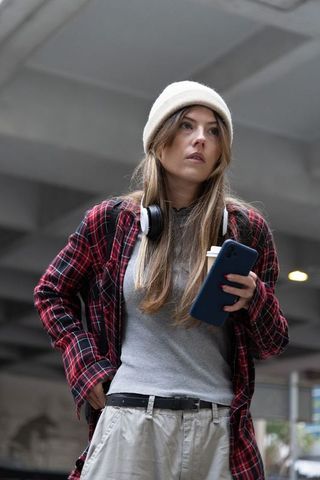 Young woman standing in parking garage with smartphone and coffee, wearing beanie and plaid