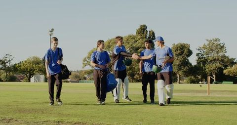 Multiethnic cricket team walking off field after match, shaking hands and carrying bats