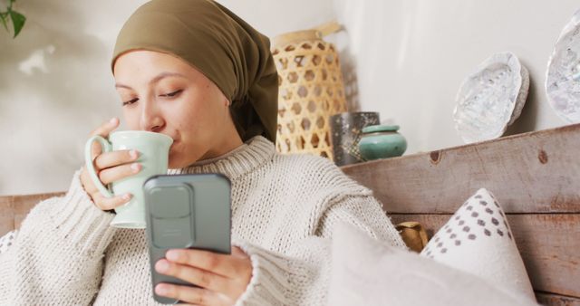 A young woman is drinking coffee while looking at her phone at home. She is wearing a cozy sweater and a headscarf. The background has a rustic and warm decor, adding to the relaxing atmosphere. This image is great for illustrating concepts of relaxation, leisure, technology usage, and lifestyle at home. Ideal for advertisements, blogs, and social media posts related to home comfort, tech, wellness, and lifestyle.