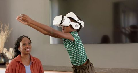 African mother smiling while watching daughter reaching and exploring with white vr headset in livin