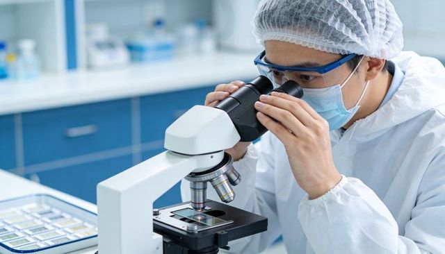 Lab technician examining microscope slide wearing protective gown goggles and face mask