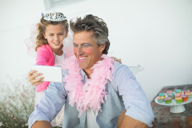 Father and daughter enjoying a playful moment in the garden. Daughter dressed in a fairy costume with a tiara, showing something on a mobile phone to her father who is wearing a pink boa. Ideal for use in family-oriented advertisements, parenting blogs, and social media posts highlighting family bonding and fun activities.
