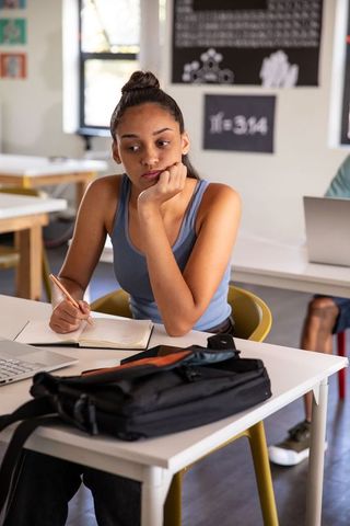 Thoughtful Teen Student Studying in Classroom with Notebook and Laptop