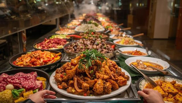 Heaping fried chicken platter at busy buffet counter with hot dishes and hands serving