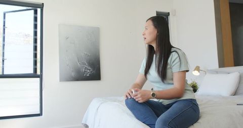 Asian woman sitting cross-legged on bed looking out window holding small white object minimalist sun