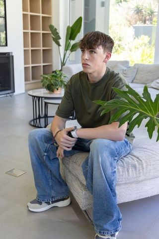 Pensive teen sitting on sofa in modern sunlit living room with casual style and greenery