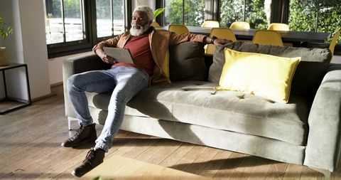 Senior man relaxing on sofa reading tablet in bright living room