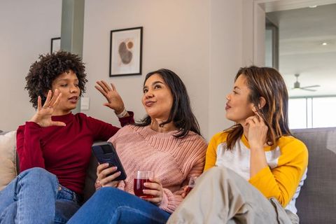 Diverse friends chatting on couch holding smartphone and red drinks, cozy home gathering