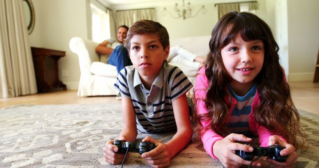 Young boy and girl are laying on carpet, engrossed in playing video games while using controllers. An adult is relaxing in the background on a couch, highlighting a cozy family home environment. Ideal for themes related to childhood, sibling bonding, indoor activities, technology engagement, family leisure time or home comfort.
