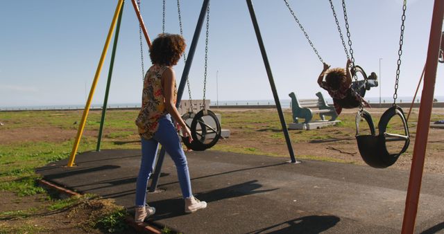 Happy biracial mother pushing son playing on swing at sunny playground. Motherhood, childhood, togetherness, summer, vacations, fun and free time, unaltered.