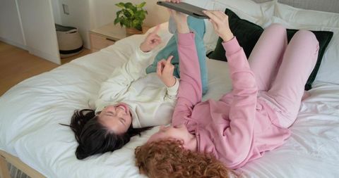 Diverse friends relaxing on bed in loungewear laughing while holding tablet