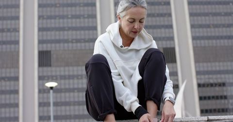Senior woman tying running shoes on urban ledge with hoodie and smartwatch