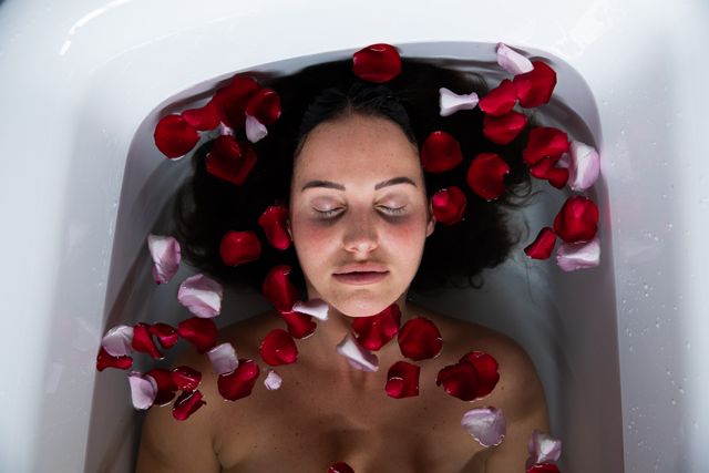 Young Caucasian brunette woman lying in a bathtub filled with rose petals, eyes closed, enjoying a moment of relaxation. Ideal for use in wellness, spa, beauty, and self-care promotions. Perfect for illustrating concepts of luxury, tranquility, and pampering.