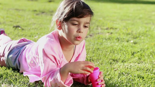 Young girl creating bubbles with a bubble wand while lying on the grass in a sunny park. Ideal for illustrating outdoor play, children's activities, summer fun, and carefree childhood moments. Suitable for family-related content, education materials on play, or advertisements related to children's products or outdoor activities.