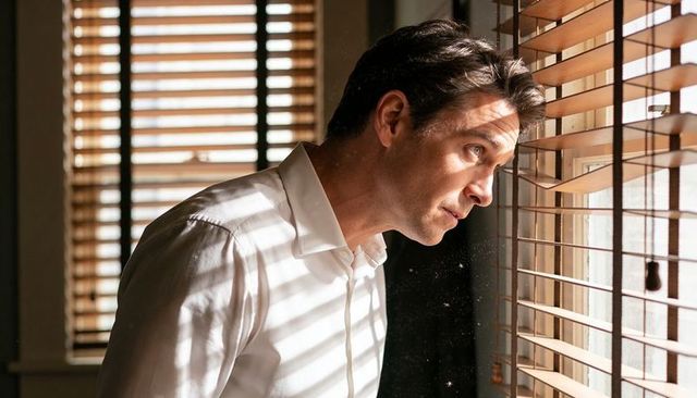 Contemplative light-skinned man peering through venetian blinds with sunlit shadows