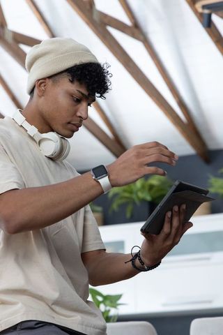 Young african american man tapping tablet in sunlit loft wearing beanie and headphones