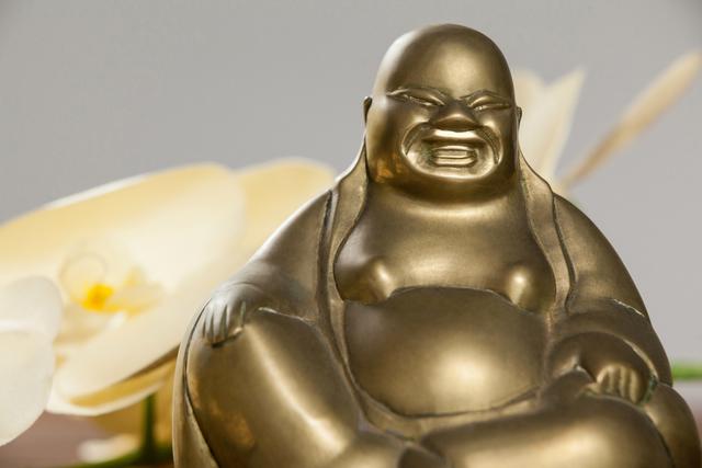 Gold Laughing Buddha figurine placed on a wooden table with white orchids in the background. Ideal for use in articles or websites related to spirituality, meditation, home decor, feng shui, and Asian culture. Perfect for promoting themes of tranquility, happiness, and prosperity.