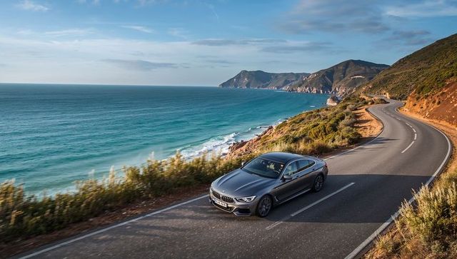 Driving silver sedan along coastal highway with turquoise sea and cliffs during golden hour